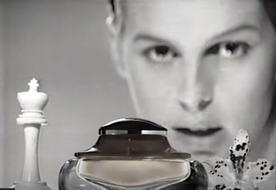 Still from Calvin Klein's 1986 Obsession commercial. black and white. Foreground from left to right: king chess piece, bottle of obsession, orchid bloom. Background: soft focus of young woman's face